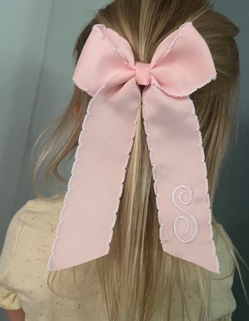 K&ouml;nnte beinhalten: Ein rosa Haarb&auml;ndchen mit einem wei&szlig;en gewellten Rand und einem wei&szlig;en gestickten Buchstaben "S".