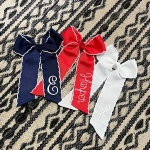 K&ouml;nnte beinhalten: Drei Haarb&auml;nder mit gewellten R&auml;ndern und personalisierter Stickerei. Die Schleifen sind marineblau, rot und wei&szlig;. Die marineblaue Schleife hat den Buchstaben "E" gestickt, die rote Schleife den Namen "Harper" gestickt und die wei&szlig;e Schleife den Buchstaben "H" gestickt.