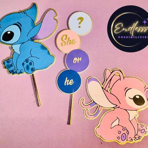 May include: A blue and pink gender reveal cake topper set featuring cartoon characters. The set includes a blue Stitch character, a pink Angel character, and signs that read "She or he?" and a question mark. The toppers have gold accents.