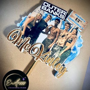 May include: A custom cake topper featuring a photo of the Outer Banks cast. The topper includes the text "Outer Banks", "Happy 13th Birthday", and the name "McKinley" in gold glitter. The base has a gold stem and a black circle with the text "Endlesss Possibilities".