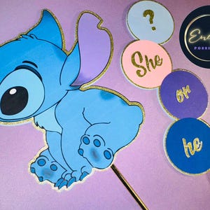 May include: A blue cartoon character with gold glitter trim, along with gender reveal party decorations. The decorations include circular signs with the words "She" and "He" in gold lettering, and a question mark.
