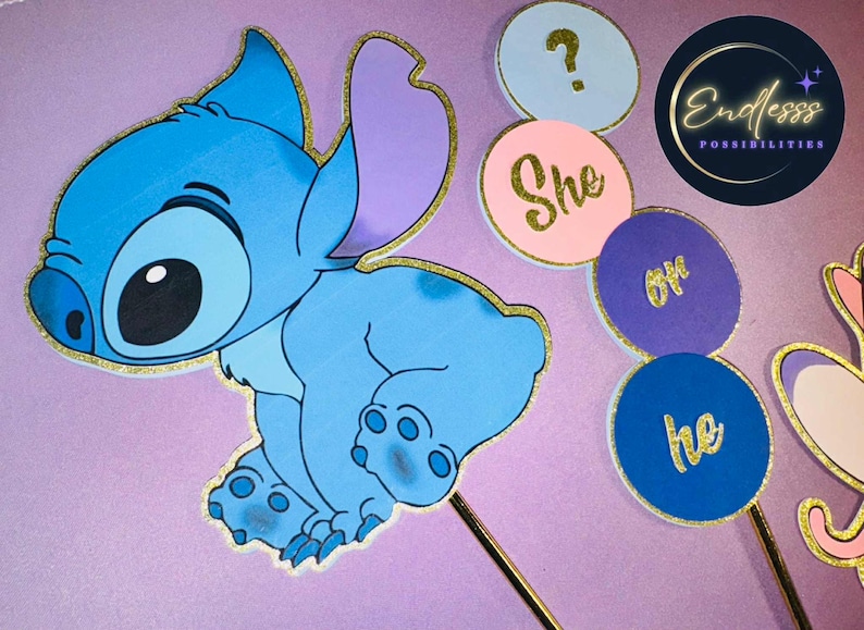 May include: A blue cartoon character with gold glitter trim, along with gender reveal party decorations. The decorations include circular signs with the words "She" and "He" in gold lettering, and a question mark.