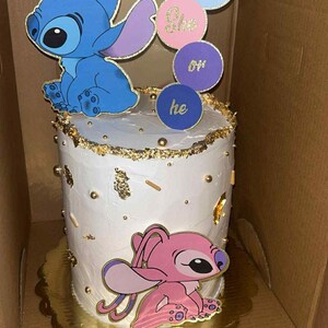 May include: A white cake decorated with blue and pink cartoon characters, gold accents, and the words "She" and "He" on circular toppers. The cake is on a gold-colored cake board.