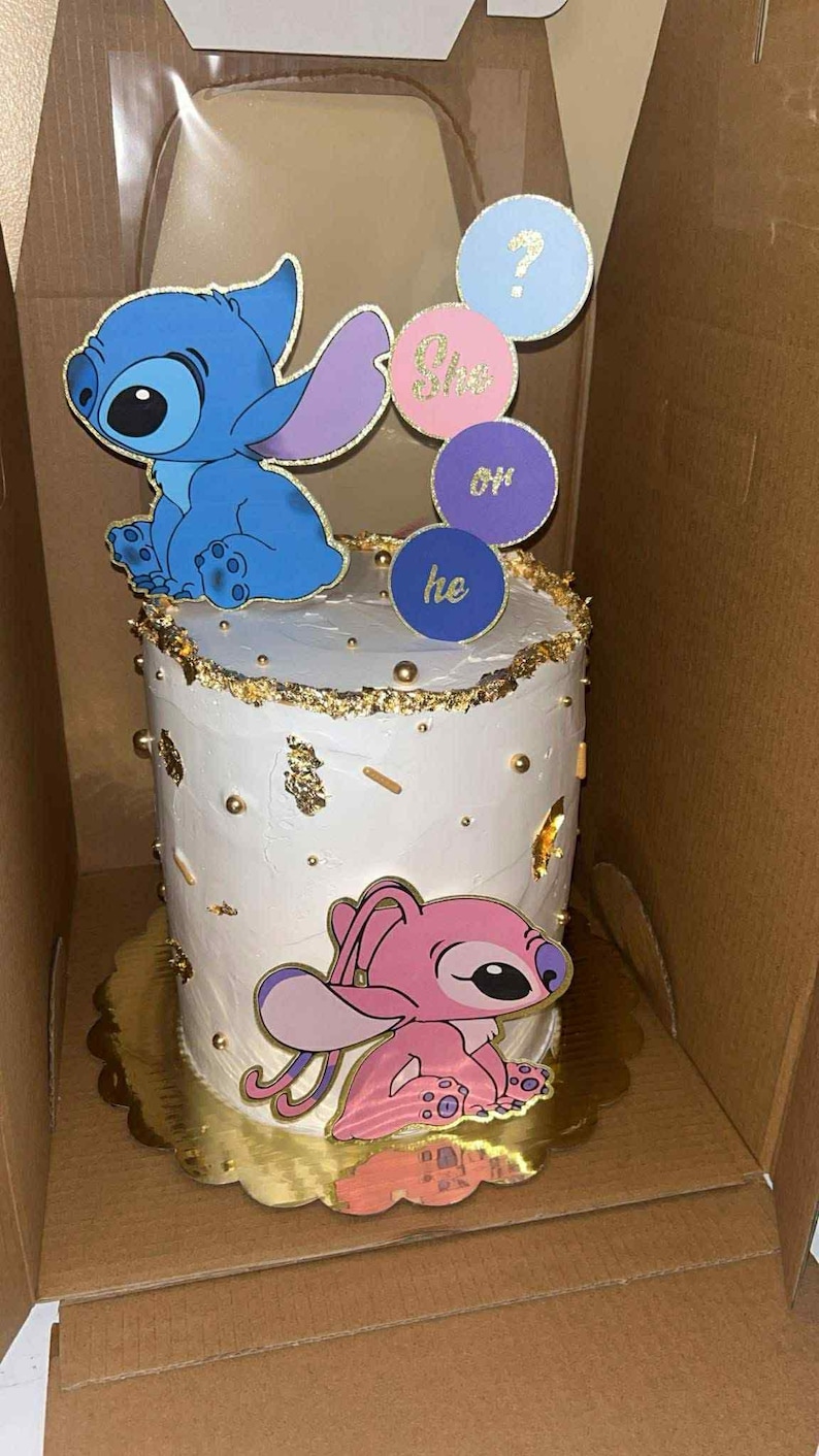 May include: A white cake decorated with blue and pink cartoon characters, gold accents, and the words "She" and "He" on circular toppers. The cake is on a gold-colored cake board.
