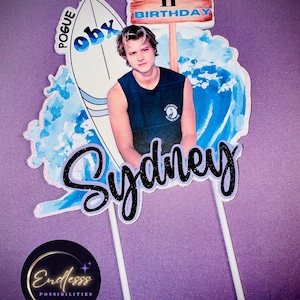 May include: A cake topper featuring a cartoon image of a young man with brown hair, wearing a black tank top, standing on a surfboard with the words "OBX" and "POGUE" written on it. The surfboard is in front of a blue and white watercolor wave. A wooden sign above the man reads "HAPPY 11TH BIRTHDAY". The words "Sydney" are written in black and white glitter letters below the surfboard.