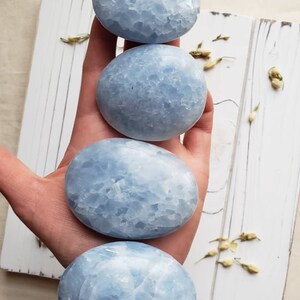 May include: Four smooth, light blue celestite palm stones, each with a unique, textured surface. The stones are stacked on top of each other, with the largest stone at the bottom and the smallest stone at the top.