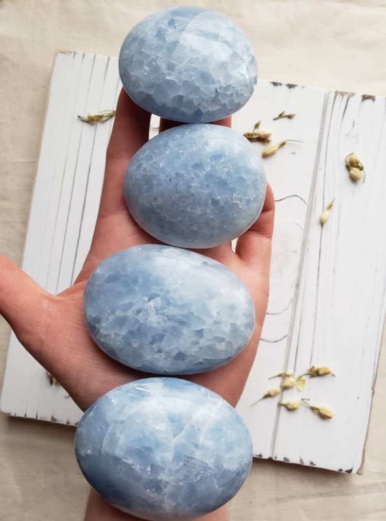 May include: Four smooth, light blue celestite palm stones, each with a unique, textured surface. The stones are stacked on top of each other, with the largest stone at the bottom and the smallest stone at the top.