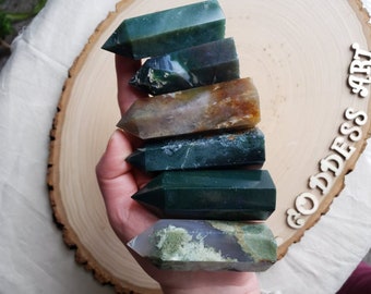 May include: A collection of seven green, brown, and grey agate crystal points, stacked on top of each other in a hand. The points are polished and have a smooth, glassy finish. The background is a wooden surface with the text "Goddess Art" written on it.