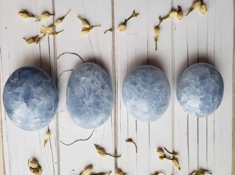 May include: Four smooth, light blue celestite palm stones arranged in a row on a white wooden surface. The stones have a cloudy, marbled appearance.