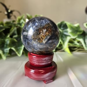 Può includere: Una gemma sferica lucidata con inclusioni blu e marroni, esposta su un supporto di legno rosso lucido. La superficie della sfera riflette la luce, mostrando i suoi intricati motivi. Il supporto ha una base rotonda e un design a gradini.