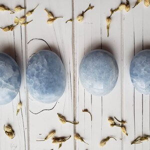 May include: Four smooth, light blue, polished, round, celestite stones on a white wooden background.