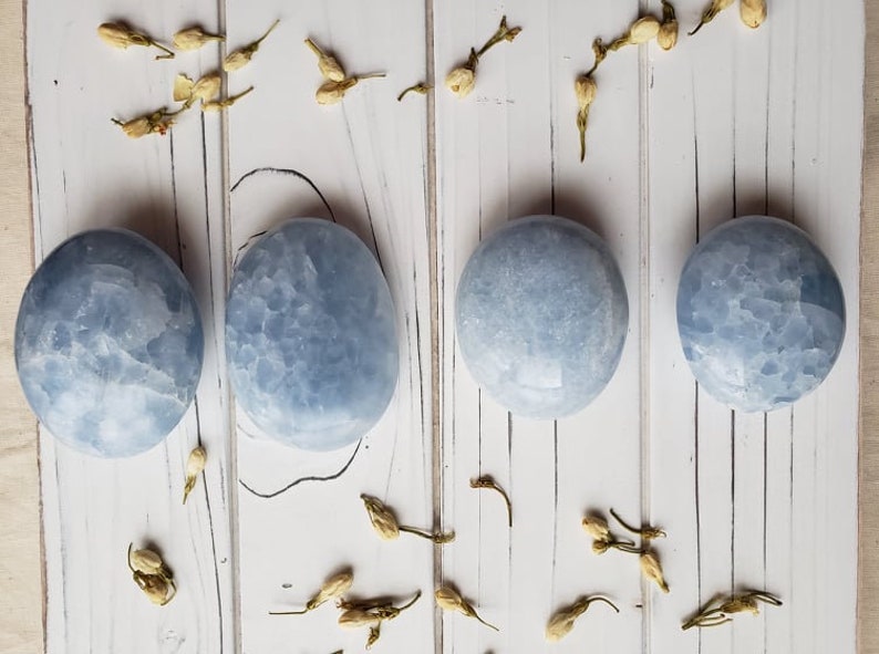 May include: Four smooth, light blue, polished, round, celestite stones on a white wooden background.