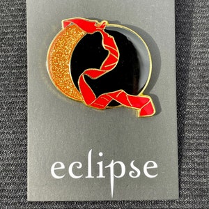 May include: A gold-colored pin with a black crescent moon and a red ribbon. The crescent moon has a glittery texture. The ribbon is tied around the moon and has a red and black striped pattern. The pin says "eclipse" in white text below the image.