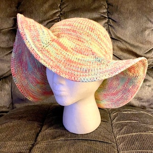 Peut inclure: Un chapeau de soleil à large bord avec un motif rayé rose, bleu et jaune. Le chapeau est crocheté et a un bord souple.
