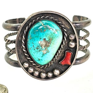 May include: A silver cuff bracelet with a turquoise stone set in a circular design. The bracelet has a rope-like design on the band and a small red stone accent.
