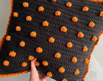 Puede incluir: Un cojín de ganchillo negro con detalles de calabazas naranjas. El cojín tiene un borde naranja y forma de estrella. Las calabazas están distribuidas uniformemente por la superficie, creando un tema festivo de Halloween.