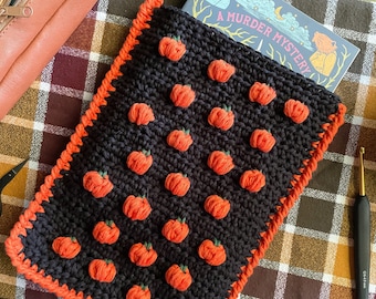 May include: A black crochet book cover with an orange border and a pattern of orange pumpkins. The book cover is holding a book titled "A Murder Mystery".