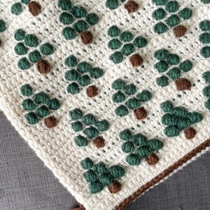 May include: A white crocheted blanket with a repeating pattern of green and brown trees. The blanket has a brown crocheted border.