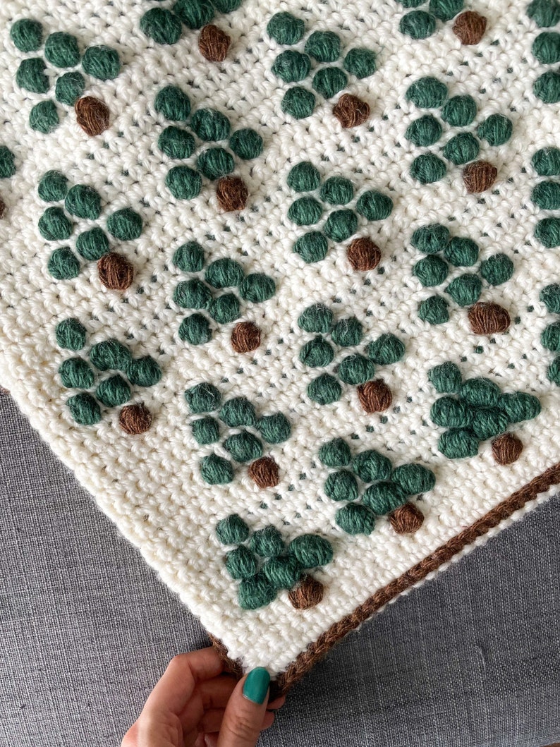 May include: A white crocheted blanket with a repeating pattern of green and brown trees. The blanket has a brown crocheted border.