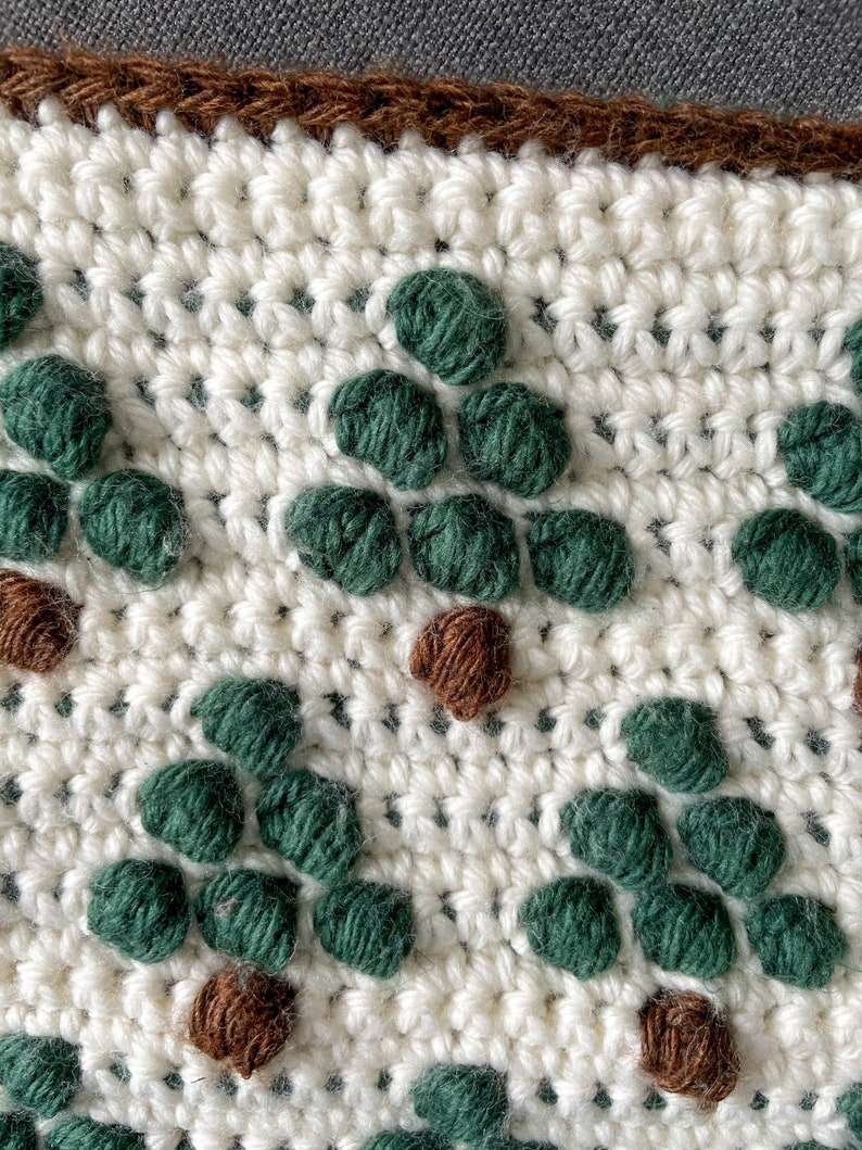 May include: Close-up of a crocheted fabric with a repeating pattern of green and brown yarn circles. The fabric is white and has a brown border.