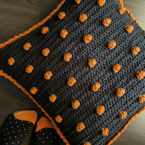 Pu&ograve; includere: Un cuscino quadrato all'uncinetto blu scuro con accenti di zucche arancioni. Le zucche sono distribuite uniformemente sulla superficie. Il cuscino ha un bordo arancione, creando un tema autunnale.