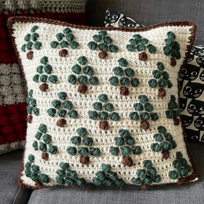 May include: A white crocheted pillow with a brown border and a repeating pattern of green trees. The trees are made with a textured stitch and have brown centers.