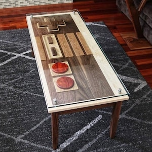 Op de afbeelding: Een houten salontafel in de vorm van een vintage videogamecontroller. De tafel heeft een glazen blad en is gemaakt van walnoot hout. De tafel heeft twee rode knoppen en een directionele pad.