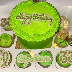 Puede incluir: Un vibrante pastel de cumpleaños verde y cupcakes en una caja blanca. El pastel está decorado con un adorno dorado y rosa que dice "Happy Birthday". Los cupcakes tienen adornos decorativos con números y signos del zodiaco.