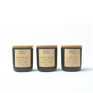 May include: Three dark gray candles with cork lids are displayed. Each candle has a label with text, including "VELVET NOCTURNE", "NATURALLY BEESWAX", and "ISLAND LUXE". The labels also mention "Beeswax & Coconut Oil Blend" and "Essential Oils".