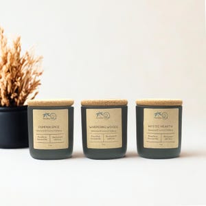 May include: Three matte dark grey candle jars with light brown cork lids and tan labels. The labels feature the text Pumpkin Spice, Whispering Woods, and Mystic Hearth, along with the phrase Beewax & Coconut Oil Blend and 5 oz.