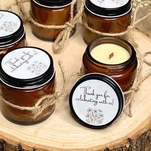 May include: Set of small amber glass jars with lids and twine tied around them. Each jar contains a white candle with a wooden wick. The lids have round white labels with brown floral designs and the text "Thank you for celebrating with us!"