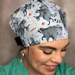 May include: A light blue surgical scrub cap featuring a repeating pattern of raccoons, trash cans, and food items. The raccoons are depicted in gray and black, with the trash cans in gray and white. The cap is tied in the back with a black tie.