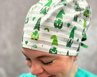 May include: A scrub cap featuring a pattern of green gnomes holding shamrocks and small rainbows against a background of light gray and white horizontal stripes. The cap includes a tie-back closure with green ribbons visible at the side.