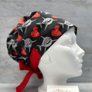May include: Black scrub cap with a red tie. The cap has a repeating pattern of white sharks with red hearts that say "Love Bites".