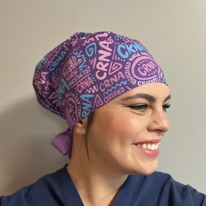 May include: A purple scrub cap with the text "CRNA", "CERTIFIED", "REGISTERED NURSE ANESTHETIST" in pink and light blue. The cap is tied in the back with a matching purple ribbon. The person is wearing a navy blue scrub top.