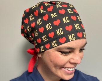 May include: A black scrub hat with a red tie. The hat has a repeating pattern of red hearts and the letters "KC" in gold.
