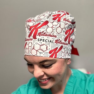 K&ouml;nnte beinhalten: Eine wei&szlig;e chirurgische M&uuml;tze mit einem roten und schwarzen Aufdruck des Wortes "Ketamin" und einer chemischen Struktur. Die M&uuml;tze hat ein rotes und schwarzes Punkte-Muster.
