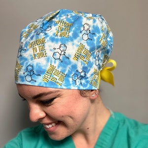 May include: A blue and white tie-dye surgical scrub cap with the text "WELCOME TO THE K HOLE" in yellow. The cap features a repeating pattern of chemical structures. A yellow ribbon is tied on the back.