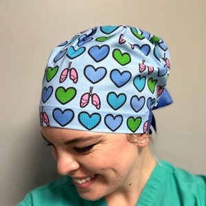 May include: A light blue surgical scrub hat with a pattern of green and blue hearts and pink and white lungs. The hat is tied at the back with a blue ribbon.