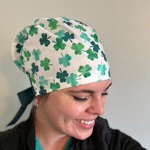 May include: A white scrub cap with a green shamrock pattern. The cap has a tie at the back.