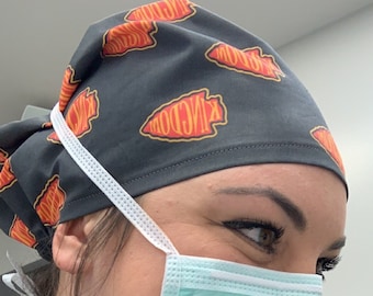 May include: A gray scrub cap with a repeating pattern of red and yellow arrowheads and the word "Kingdom". A light blue surgical mask is worn with white straps. The person is wearing a blue scrub top and a black jacket.