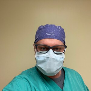 May include: A person wearing a blue surgical cap with the text "Sweet dreams are made of these" and a light blue surgical mask. The person is also wearing a green surgical scrub top.