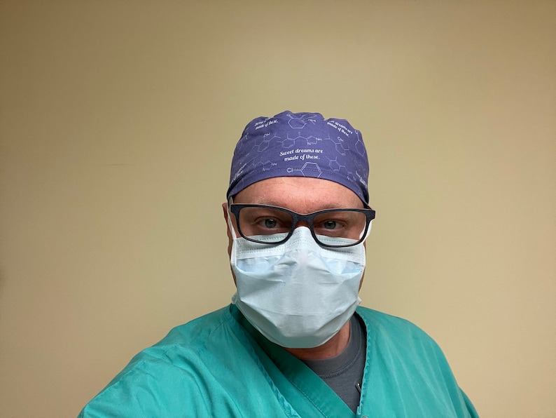May include: A person wearing a blue surgical cap with the text "Sweet dreams are made of these" and a light blue surgical mask. The person is also wearing a green surgical scrub top.