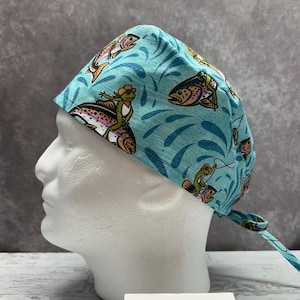 May include: A teal scrub cap featuring a whimsical pattern of fish and frogs. The fish are depicted with colorful details, and the frogs are riding the fish. The cap has a tie in the back and is displayed on a mannequin head.