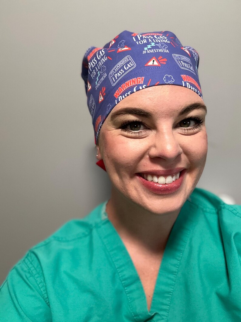 May include: A blue scrub hat with a white and red pattern. The hat has the text "I Pass Gas For A Living #Anesthesia" printed on it.
