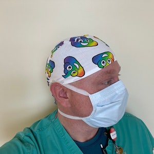 May include: A white surgical cap with a repeating pattern of rainbow-colored smiling poop emojis. The person is also wearing a light blue surgical mask and green scrubs.