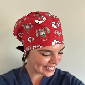 May include: Red surgical scrub cap with a repeating pattern of a cartoon wolf in a football jersey and the Kansas City Chiefs logo. The cap has a black tie in the back.