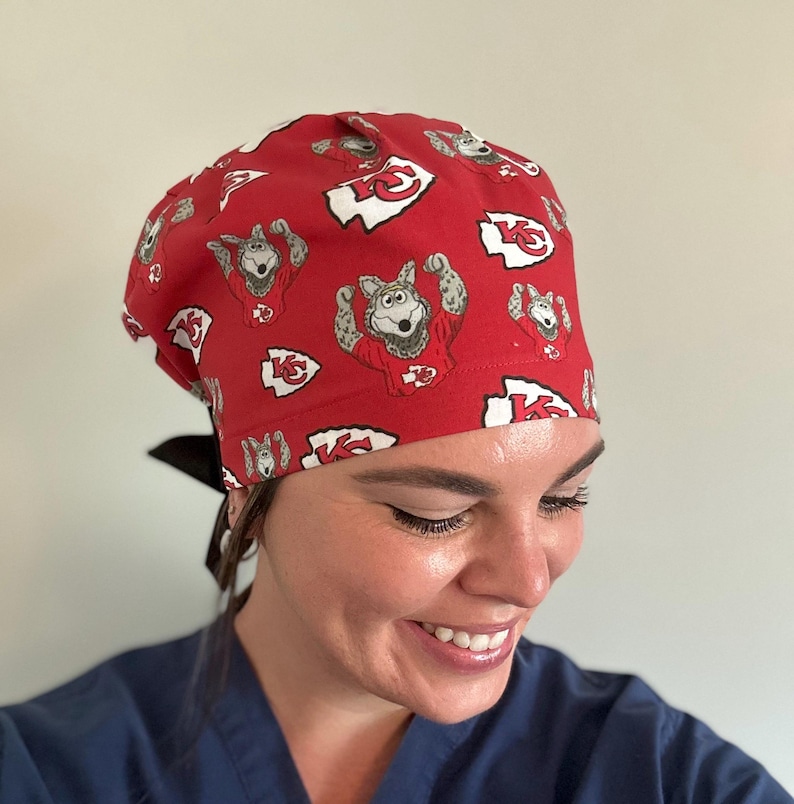May include: Red surgical scrub cap with a repeating pattern of a cartoon wolf in a football jersey and the Kansas City Chiefs logo. The cap has a black tie in the back.