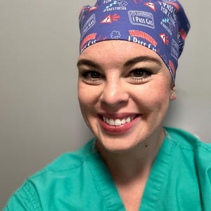 May include: A woman wearing a blue scrub cap with a white and red print that says "I Pass Gas For A Living #Anesthesia". She is smiling and wearing a teal scrub top.