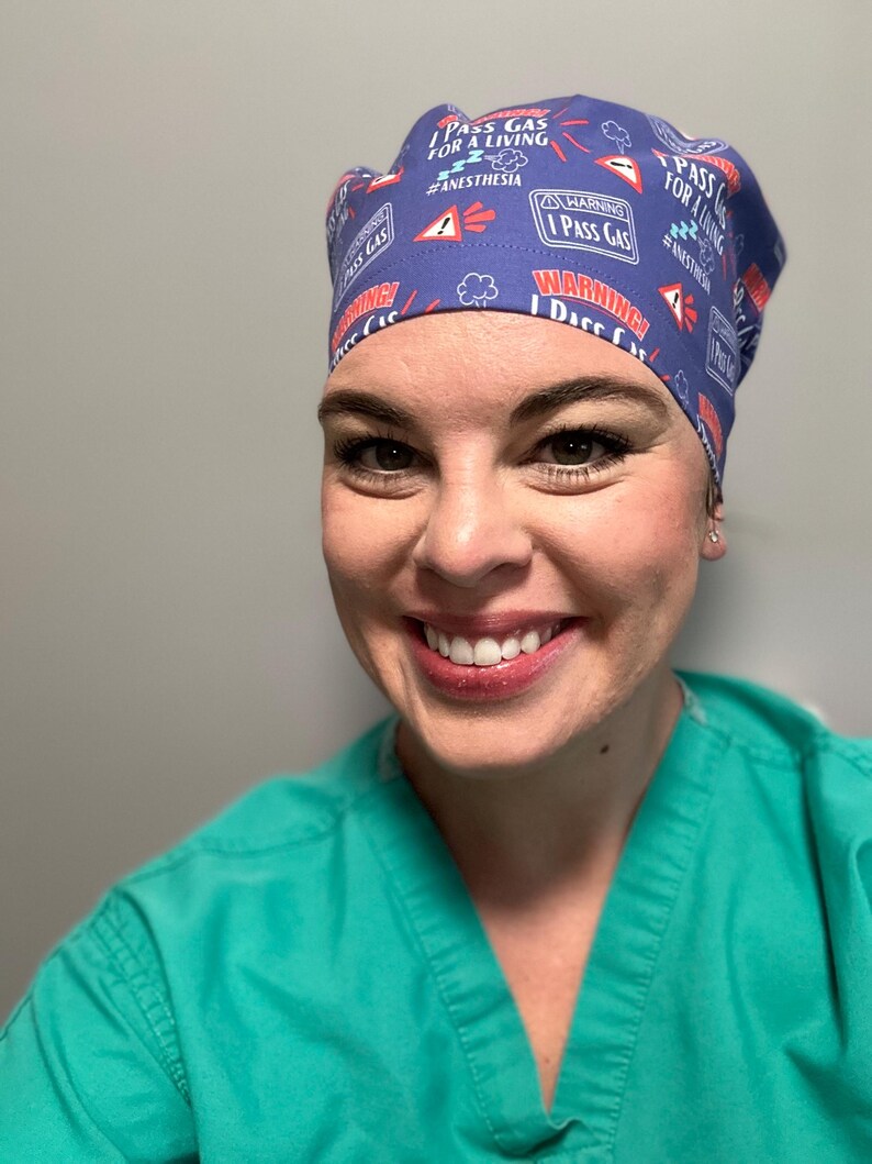 May include: A woman wearing a blue scrub cap with a white and red print that says "I Pass Gas For A Living #Anesthesia". She is smiling and wearing a teal scrub top.